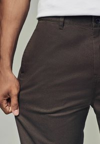 Pantalons marron foncé avec une texture lisse, présentant une poche avant, des passants de ceinture et une fermeture à un bouton au niveau de la taille.