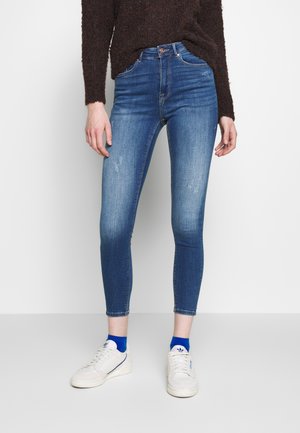 Jeans ajustados en denim azul medio, con una cintura alta, ligero desvanecimiento y un ajuste ceñido. Combinados con zapatillas blancas y calcetines azules.