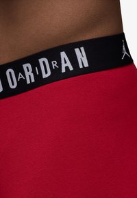 Røde sportsshorts med en glat tekstur og en bred sort talje, der har hvid tekst "JORDAN" og et logo.