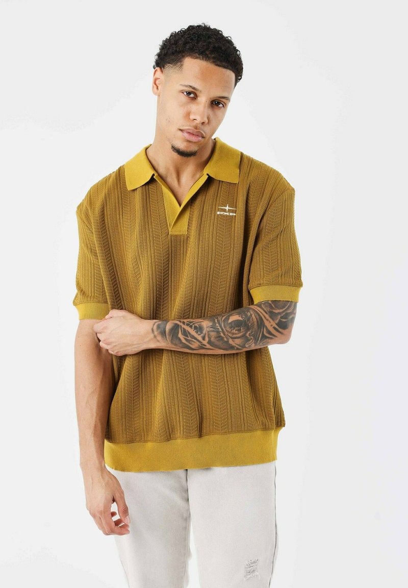 Polo shirt tricot jaune moutarde avec des rayures texturées, manches courtes et ourlet côtelé. Présente un logo discret sur la poitrine.