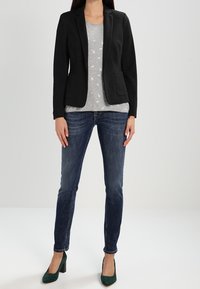 Blazer negro con dos bolsillos frontales sobre una camiseta gris con estampados de animales blancos, combinado con jeans azul oscuro y tacones verdes.