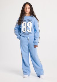 Girl with long curly hair wearing a light blue tracksuit with "New York 89" text and white sneakers, standing against a plain background.