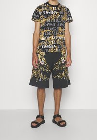 Svart kortärmad skjorta med guld- och vit logotypdesign; svarta shorts med utsmyckad flerfärgad broderi; ihop med svarta sandaler.