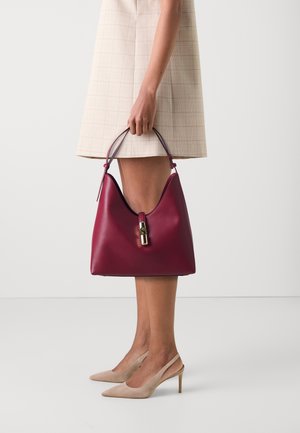Sac en cuir bordeaux avec une silhouette incurvée, une poignée supérieure et des détails en métal doré. Porté avec une robe à carreaux et des talons aiguilles beiges.