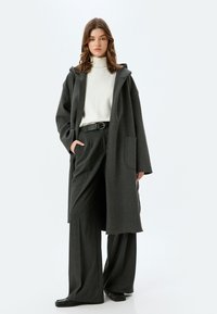 Lang, donkergrijze jas met een grote capuchon, open voorkant en zijzakken, gecombineerd met een witte coltrui en wijde donkergrijze broek.