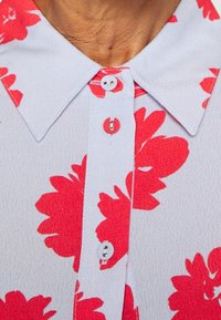 Zbliżenie na jasnoniebieską koszulę z czerwonym kwiecistym wzorem i trzema widocznymi guzikami na przedniej listwie pod kołnierzem.