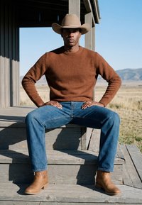 Homme portant un pull marron, un jean bleu, des bottes de cowboy couleur sable et un chapeau de cowboy couleur sable, assis sur des marches en bois en plein air dans un paysage rural.