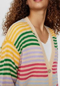 Žena s kudrnatými vlasy oblečená v pruhovaném kardiganu v mnoha barvách přes bílý top, který odhaluje linii dekoltu a detaily knoflíků.