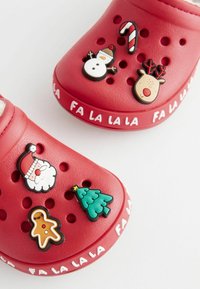 Röda träskor med flera dekorativa 3D-charms: jultomte, snögubbe, godiskäpp, ren, gran och pepparkaksgubbe. "FA LA LA LA LA" text på sidan.