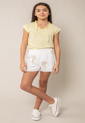 Jeune fille debout, jambes croisées, portant un haut jaune à manches courtes, un short blanc à imprimé palmiers, et des baskets blanches.
