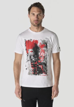 Hombre con camiseta gráfica blanca con diseño abstracto en rojo, negro y gris, de pie frente a un fondo liso gris claro.