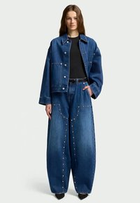 Jovem mulher vestindo uma jaqueta de denim azul oversized e jeans de perna larga com tachas metálicas, camisa preta e sapatos pretos de bico fino, em pé.