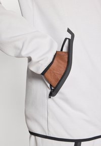 Λευκό μπουφάν με φερμουάρ και μαύρες λεπτομέρειες, με πλαϊνή τσέπη που έχει αντίθετη καφέ εσωτερική επένδυση και απαλής υφής ύφασμα.