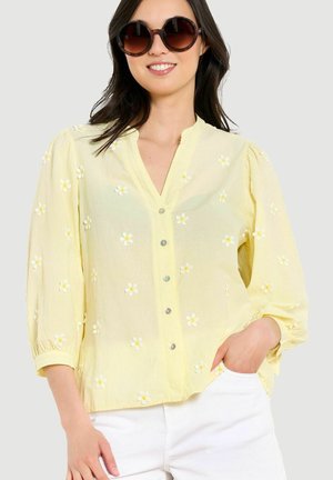 Vrouw met ronde donkere zonnebril en een lichtgele blouse met knopen en witte en gele geborduurde bloemen, poseert met één hand in de zak.