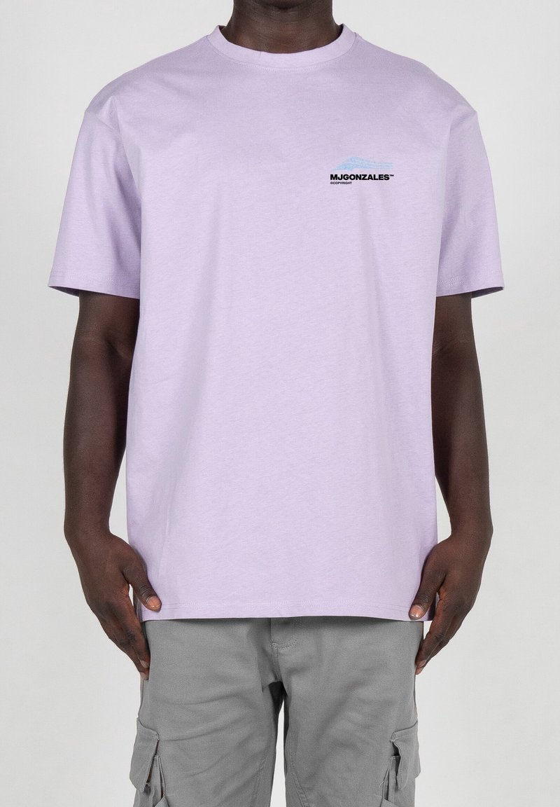 Fliederfarbenes Baumwoll-T-Shirt mit kurzen Ärmeln und rundem Ausschnitt, mit einem schwarzen Logo und einer blauen Berggrafik auf der linken Brust. Kombiniert mit grauen Cargo-Shorts.