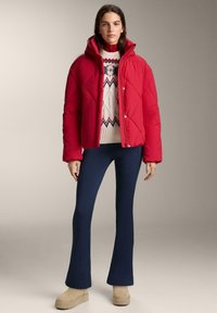 Rød pufferjakke med hætte og diamantquiltet mønster over en cremefarvet striktrøje med geometrisk design, kombineret med marineblå flared bukser.