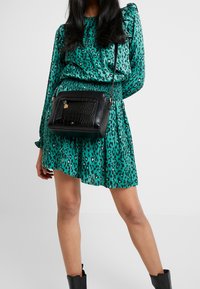 Schwarze Handtasche mit Krokodilprägung in rechteckiger Form, metallischen Details und verstellbarem Riemen. Kombiniert mit einem grünen Kleid mit Leopardenmuster.