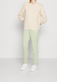Beige sweatshirt med rund halsringning och vit Nike-logotyp, kombinerad med ljusgröna joggers och vita sneakers. Mjuk tyg, avslappnad passform.