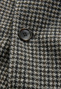 Material cu model houndstooth în nuanțe de negru, alb și maro, cu un buton mare, închis la culoare, cu patru găuri. Textură moale de lână.