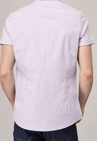 Camisa de manga corta en tela color púrpura claro, con dobladillo recto, detalle de yugo en la parte posterior y costuras laterales para un ajuste entallado.
