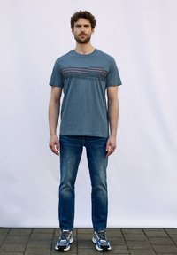 Heren blauwe katoenen t-shirt met ronde hals, horizontale streeppatroon op de bovenste borst, gecombineerd met vervaagde blauwe jeans en sneakers.