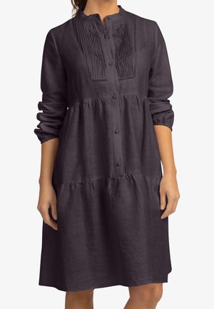 Femme portant une robe à manches longues, de couleur violet foncé, avec des boutons sur le devant, un détail plissé sur la poitrine et une taille froncée, visible du cou aux genoux.