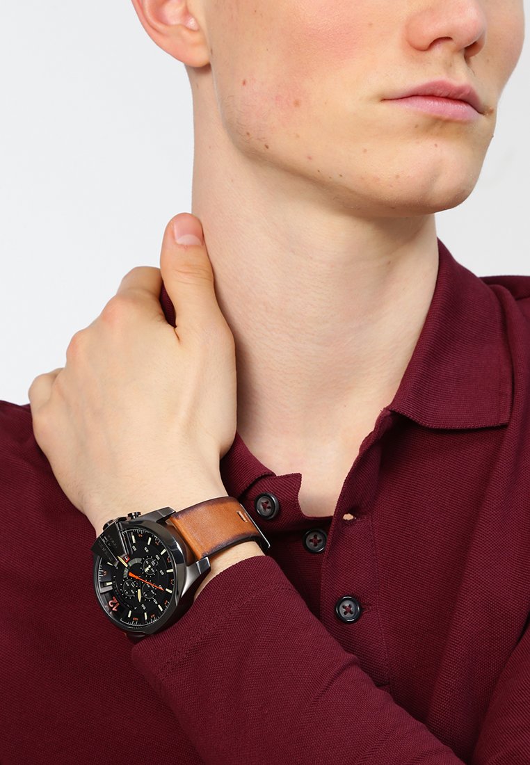Orologio nero con funzioni cronografiche, cinturino in pelle marrone, quadrante tondo con accenti rossi, indossato su un polso, abbinato a una maglietta polo bordeaux.