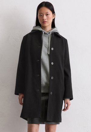 Zwarte wollen jas met een rechte snit, grote knopen en een ronde kraag, gedragen over een grijze hoodie, met een gestructureerde afwerking en een lichte glans.