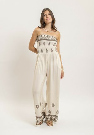 Femme debout portant une combinaison crème sans bretelles avec des motifs floraux foncés sur le corsage et les bas de pantalon, chaussée de sandales et parée de bijoux en or.