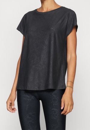 Vrouw draagt een loszittend zwart kortärmelig topje en zwarte legging met een subtiel donker patroon, staand tegen een effen lichte achtergrond.