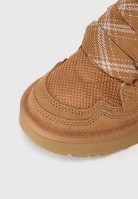Vârful sneakerilor maro cu texturi din plasă și suede, având cusături groase și un detaliu de țesătură bej cu model aproape de gleznă.