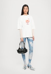 Hvit grafisk t-skjorte med havfrue-print, kombinert med blå leggings med skydesign, sølvspisse sko og en liten svart veske med nagler.