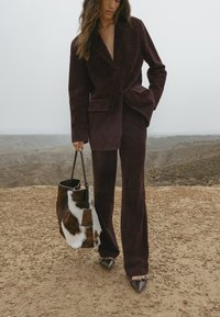 Abito in camoscio bordeaux con blazer su misura e pantaloni a gamba larga, abbinato a una borsa in pelliccia marrone e bianca e tacchi neri a punta.