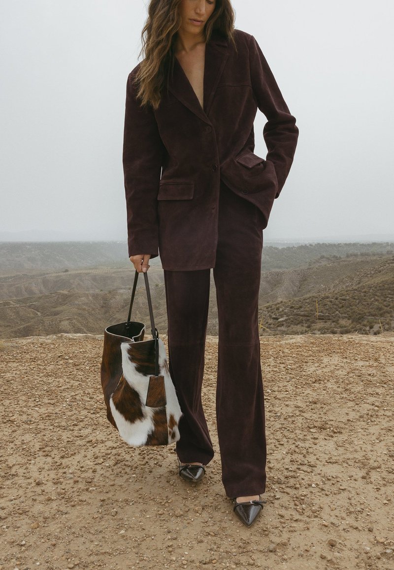 Abito in camoscio bordeaux con blazer su misura e pantaloni a gamba larga, abbinato a una borsa in pelliccia marrone e bianca e tacchi neri a punta.
