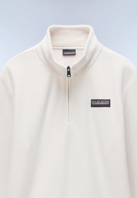 Hvid fleece-pullover med høj krave, kvart lynlås og et rektangulært logo-patch med sort og rød tekst på brystet.