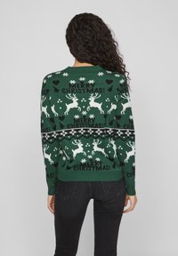 Pull de Noël vert avec des rennes blancs et des flocons de neige, avec le texte "JOYEUX NOËL !" en noir. Poignets et ourlet côtelés.