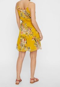 Robe fleurie jaune avec fines bretelles, présentant une taille cintrée et une jupe fluide. Les motifs incluent des fleurs colorées et des feuilles vertes.