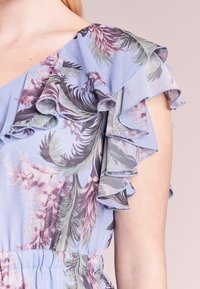 Kukallinen mekko, jossa on sininen tausta ja purppuran sekä vihreiden lehtikuviot. Rypytetyt hihat ja vyötärön kireä yksityiskohta.