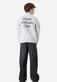 Sudadera gris con el texto y gráfico "Fresh Attitude Club" en la espalda, combinada con jeans de mezclilla oscuros que tienen grandes bolsillos.
