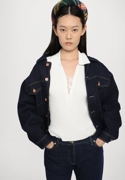 Mørk denimjakke med kobberknapper, lagdelt over en hvit, ribbestrikket topp med snøring i halsen, akkompagnert av matchende mørke jeans.