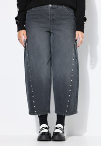 Jeans de mezclilla gris oscuro con un diseño de pierna ancha, botones a presión en los laterales y sutiles adornos. Combinados con calcetines negros y zapatos de plataforma.