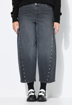 Jeans de mezclilla gris oscuro con un diseño de pierna ancha, botones a presión en los laterales y sutiles adornos. Combinados con calcetines negros y zapatos de plataforma.