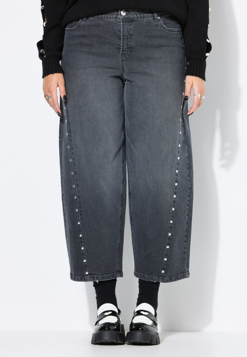 Jeans de mezclilla gris oscuro con un diseño de pierna ancha, botones a presión en los laterales y sutiles adornos. Combinados con calcetines negros y zapatos de plataforma.