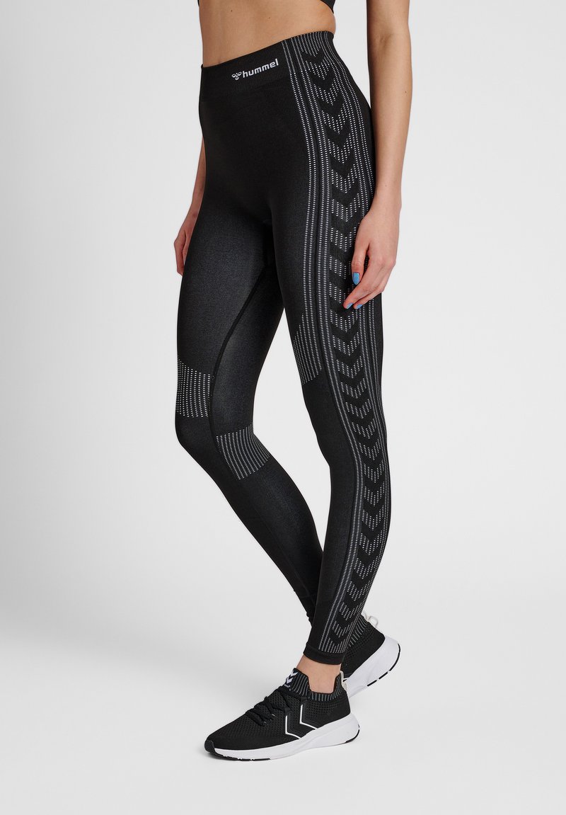 Kvinde iført sorte leggings med høj talje, med mønstrede sider og matchende sorte sneakers med hvide såler.