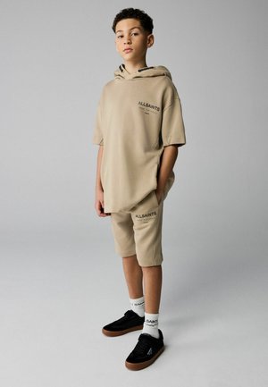 Dreng iført beige AllSaints hættetrøje og matchende shorts, sorte sneakers, hvide sokker, stående mod en ensfarvet grå baggrund.