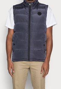 Marinblå vadderad väst med hög krage, dragkedja framtill och sidofickor. Slät yta med en rund emblem på vänster bröst.