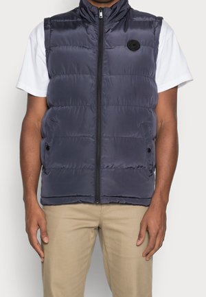 Granatowa kamizelka puchowa z wysokim kołnierzem, przednim zapięciem na zamek i bocznymi kieszeniami. Gładka tekstura z okrągłą emblematą na lewej piersi.