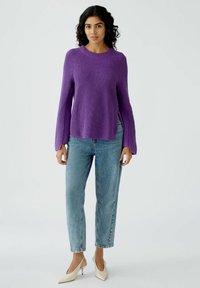 Lila gerippter Strickpullover mit langen Ärmeln, seitlichen Schlitzen, kombiniert mit hellblauen Jeans und cremefarbenen, spitzen Pumps.
