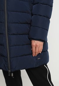 Mörkblå puffjacka med quiltade sektioner, dragkedja fram, och märkesdetalj. Mjuk textur, sidofickor och långa ärmar.