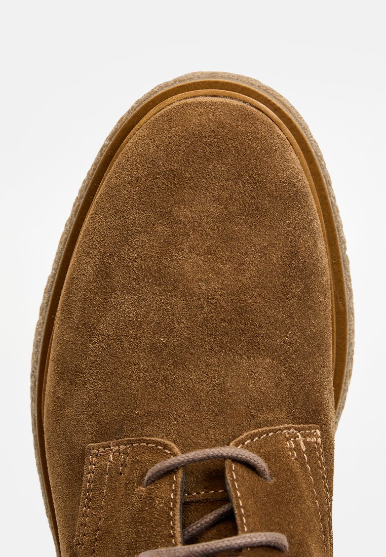 Braune Wildleder-Schuhe mit runder Zehenpartie, sichtbaren Nähten und braunen Schnürsenkeln, aus einer Nahaufnahme von oben gezeigt.